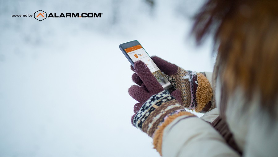 A woman in a cozy coat and gloves checks on her Alarm.com app on her phone while standing in the snow.