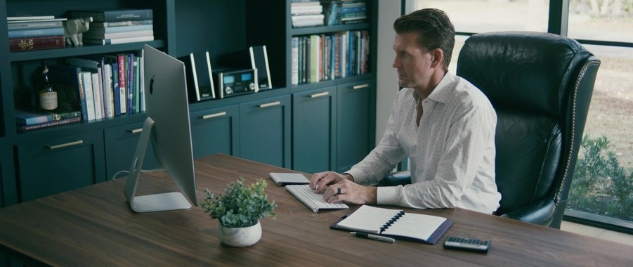 man sitting in a home office using an imac computer. There is a plant on the desk and bookshelves in the background. 