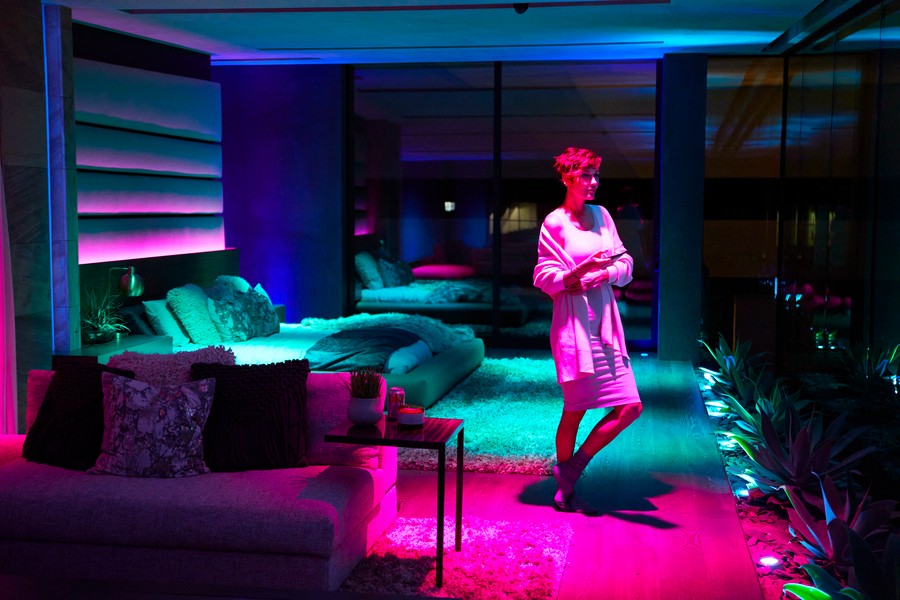 A woman in a bedroom lit by magenta, pink, teal, and purple lighting.