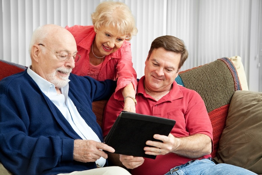  Two men on a couch holding a tablet and smiling with a woman behind the couch pointing at something on the tablet screen.