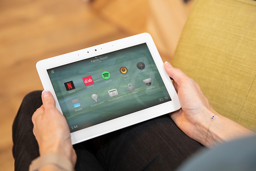 A person holds a white tablet displaying a Control4 smart home interface labeled "Family Room," featuring options for Netflix, Spotify, lighting, security, and more. 