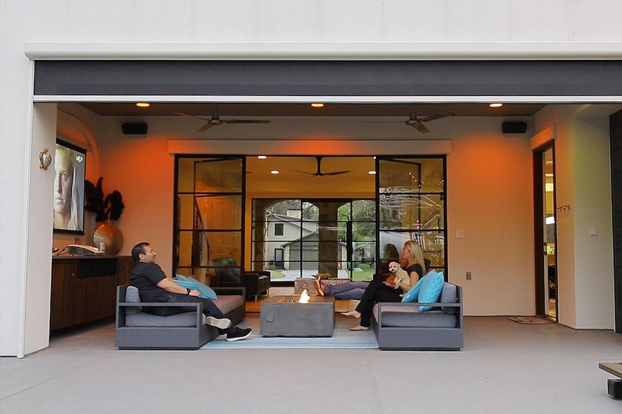 A couple relaxes on an outdoor patio equipped with sofas, an outdoor TV, and a large motorized shade.