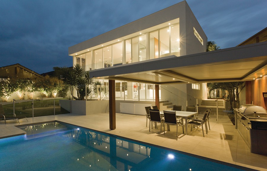 outdoor view of a pool area, seating, and a luxury home illuminated from the inside and out.